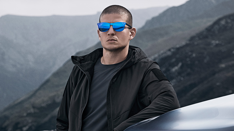 A male model leans against the bonnet of a Defender vehicle. He wears squared frames with a highly mirrored frame front in a metallic blue. His clothes are black and look suitable for a harsh outdoor environment. In the background, rugged dark hills fade into the background