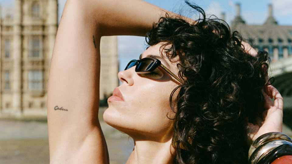 A model poses, scrunching her curly black hair, on a boat in front of Big Ben. It is sunny and bright with blue skies and she wears tiny black sunglasses