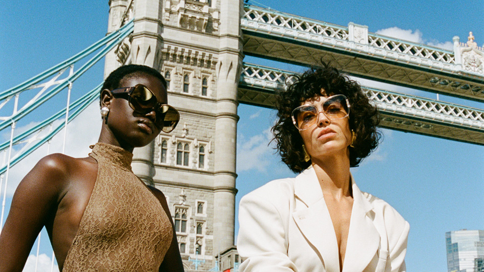 Two models pose in sunglasses on a boat along the Thames, behind them is Big Ben