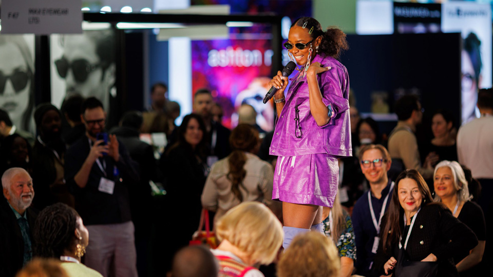 A presenter stands on a central catwalk stage speaking with the crowd. She is wearing bright purple-pink shorts and matching oversized jacket. She wears dark oval sunglasses with a pastel chunky glasses chain