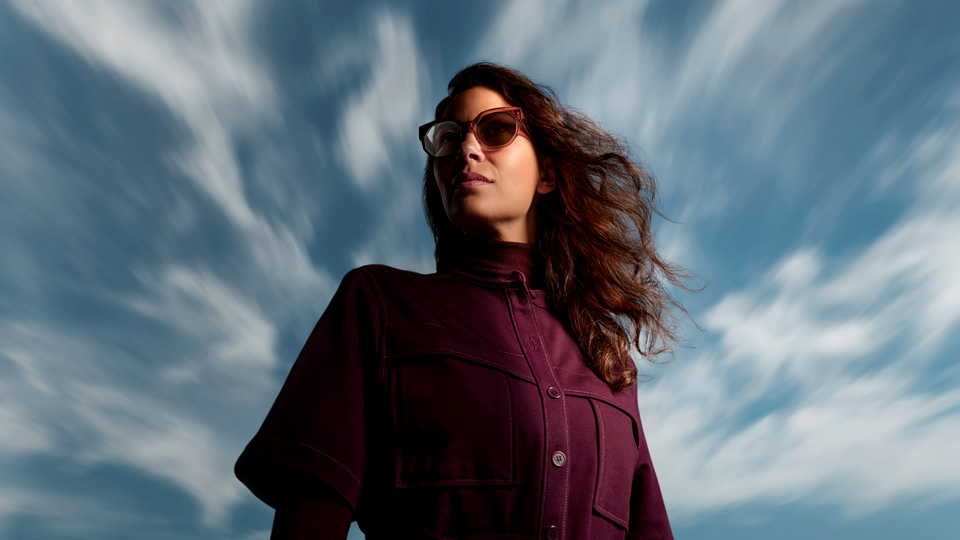 A woman wearing a plum coat and matching cat’s eye style frames stands against a bright blue sky with wispy white clouds. One lens in her frames is clear while the other is darkened