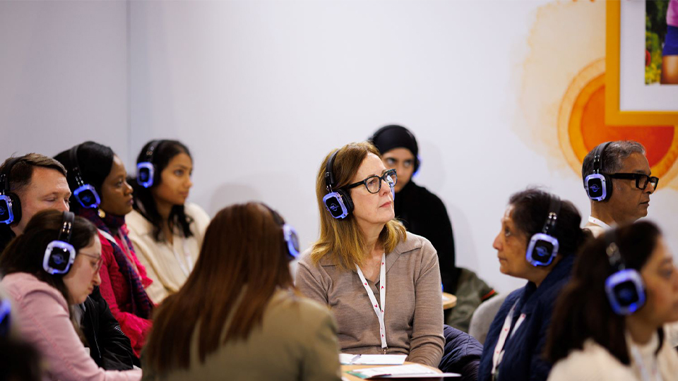 A group sitting at circular tables are all turned to listen to a presentation. They all wear headphones which glow purple-blue around the edge