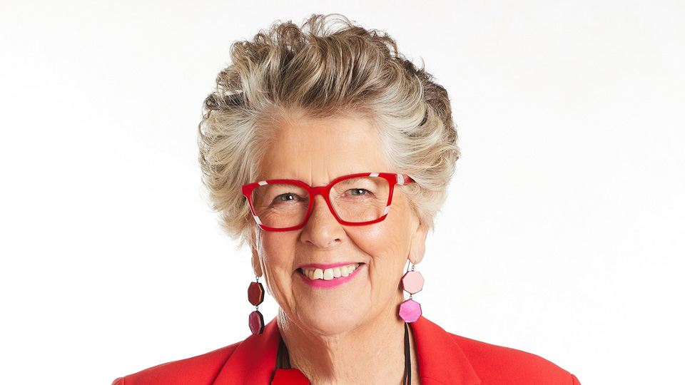 Prue Leith stands confidently smiling. She wears a pair of squared spectacles with a bright ruby red frame decorated with slices of white-grey acetate. She wears a matching red blazer and a pink, red, black and white necklace