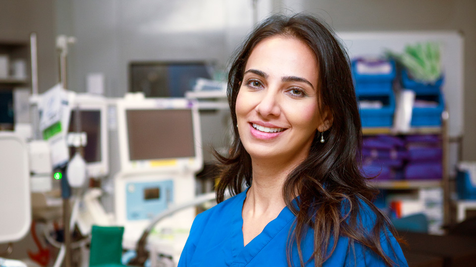 Sana sits in a clinical operating room wearing cobalt blue scrubs with her shoulder-length brown hair down. Behind her is the equipment of an ophthalmic operating room including a lot of machines and computers
