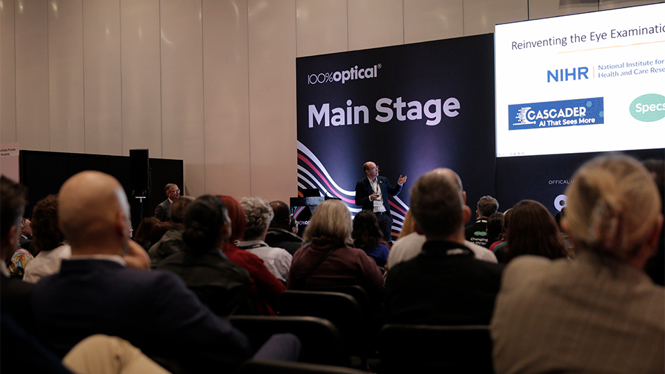Rows of attentively listening audiences look towards the stage where a man presents on the new company Cascader. The stage is branded as 100 percent optical