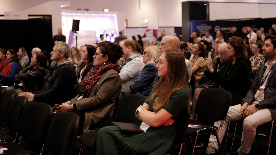 An audience watches a presentation at 100% Optical