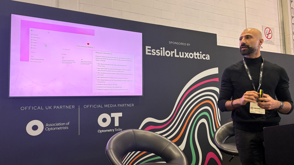 Kishan Devraj, wearing a black outfit and conference lanyard, speaks on a small stage, holding a presentation clicker. Behind him is a dark branded backdrop reading “Sponsored by EssilorLuxottica,” with logos for the Association of Optometrists and Optometry Today. A large screen displays a slide with a pink interface, and two black curved chairs sit in front of the backdrop