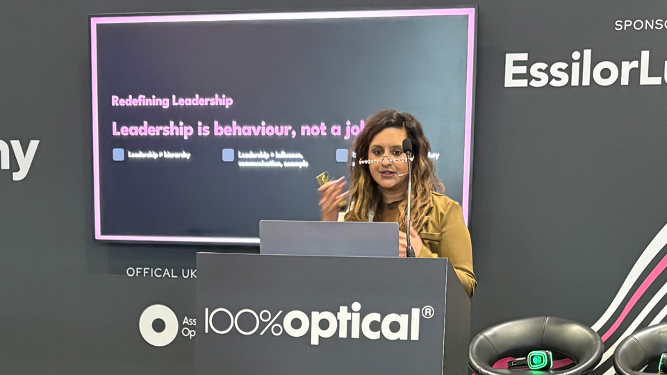 A woman speaks at a podium on stage at 100% Optical, delivering a presentation titled “Redefining Leadership.” A screen behind her displays the message “Leadership is behaviour, not a job,” while branded signage and stools sit to the side