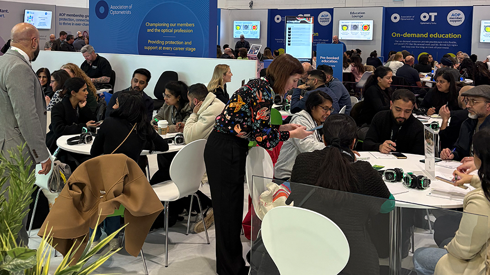 Busy seating area at a professional exhibition with a large sign reading “AOP Lounge & Café.” Groups of attendees sit at round tables in discussion while others stand nearby, with presentation screens and optometry-related displays visible in the background