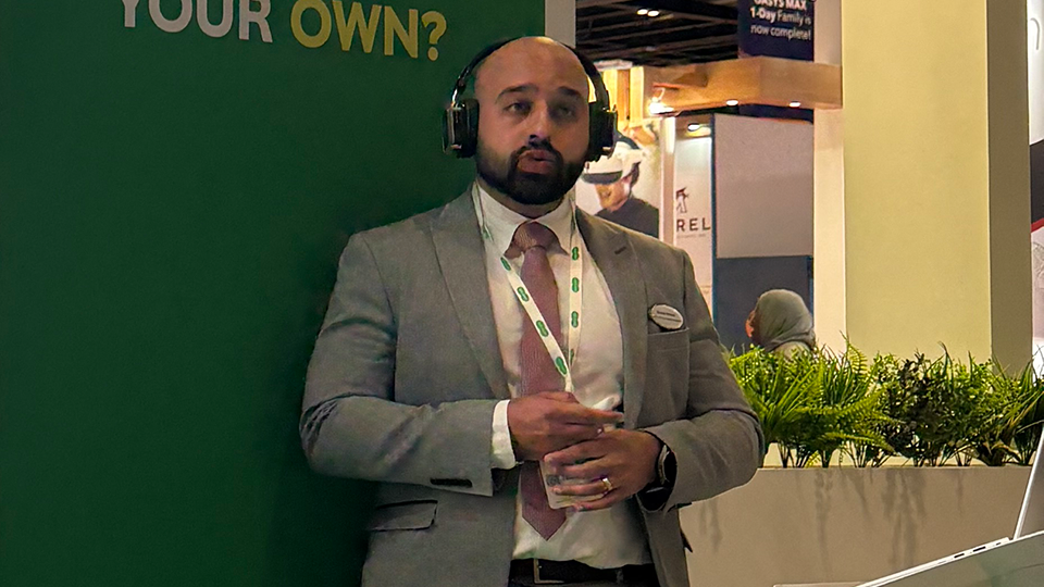A speaker wearing a grey suit and headset microphone, Bhavik Parmar, stands beside a large presentation screen at an exhibition stand with green branding. Text on the wall reads “Want to change lives including your own?” while a slide referencing NICE glaucoma guidance is partially visible on the screen