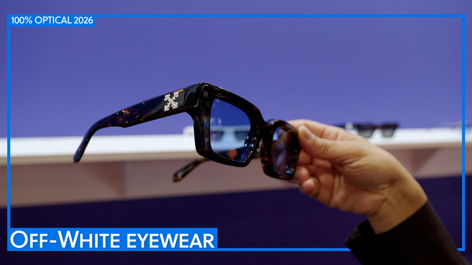A hand holds a pair of bold glasses up to the camera. The glasses are a thick chunky brown acetate in a squared style with light blue tinted lenses. On the arm of the frame is a large white logo of two double-headed arrows crossed
