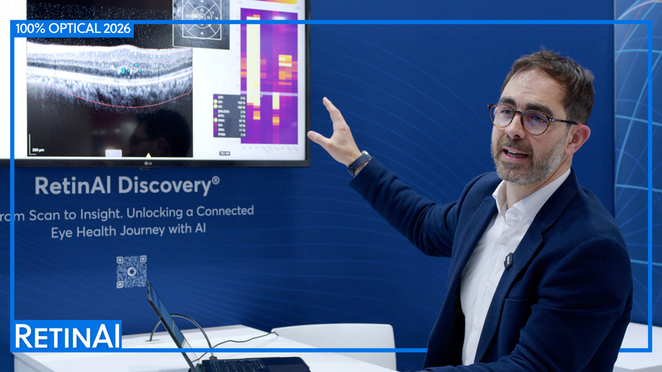 Dr Carlos Ciller, in a navy suit jacket and white shirt, is seated in the RetinAI exhibition stand at 100% Optical. He gestures at a screen behind him which displays the company’s platform