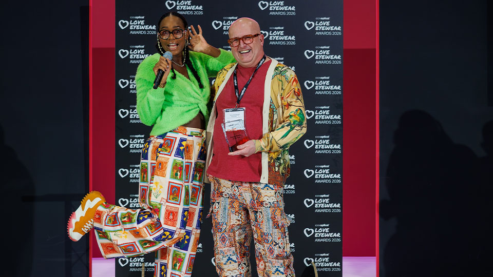 Billy Brown wears a colourful outfit and is handed a glass award recognising his practise as Eyewear Retailer of the Year. He stands on a colourful stage