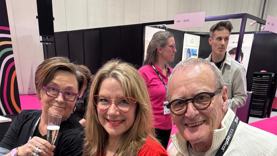 Three people are seated at an exhibition booth, they are smiling and holding up glasses