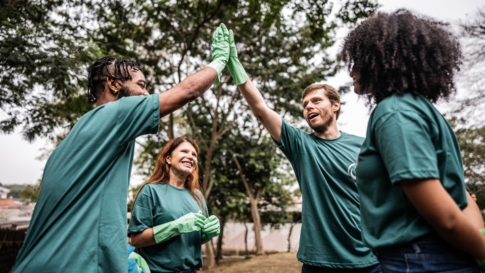 Two volunteers high five each other in a park wearing rubber gloves while other volunteers look on 