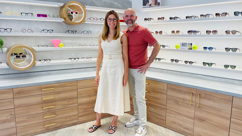 A woman and man pose in a stylish practice. The woman wears a cream-white linen dress with belt and black and white sandals, while the man wears white jeans, cream trainers, and a red polo shirt. Both wear spectacles. On the marble-effect walls around them are shelves of spectacles and sunglasses and wooden cabinets