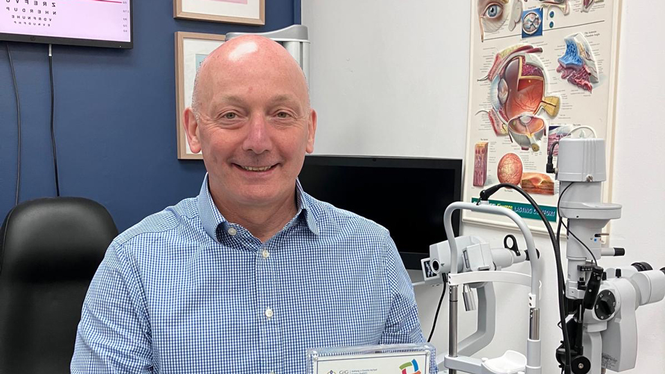 Gareth poses in an optometry practice consulting room holding a framed certificate from HEIW. Gareth wears a blue checked shirt and smiles happily. The background has one deep blue and one white wall. Clinical equipment and posters can also be seen