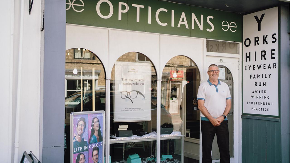Garrey, in a white polo shirt and black trousers, stands on the doorstep to an opticians. The store is painted in a sage green with arched window display. An eye chart presents the name of the company on the side