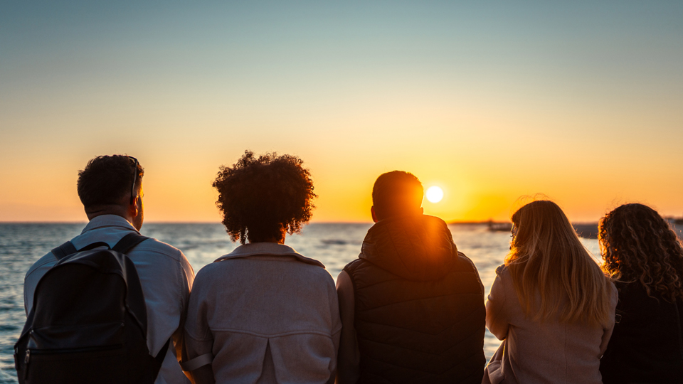 A group of friends look out at the sunset with their backs turned to the camera