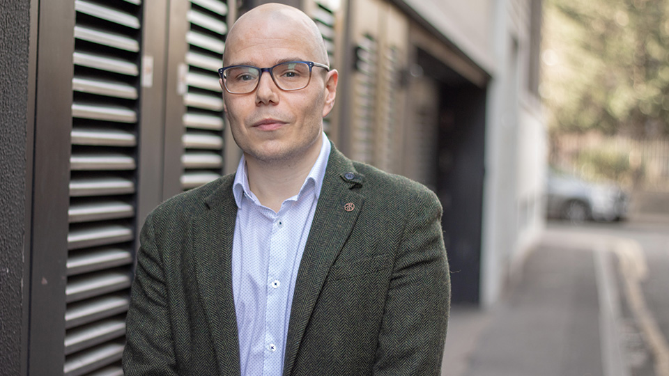 A man in a grey suit and glasses stands in front of brown doors outside