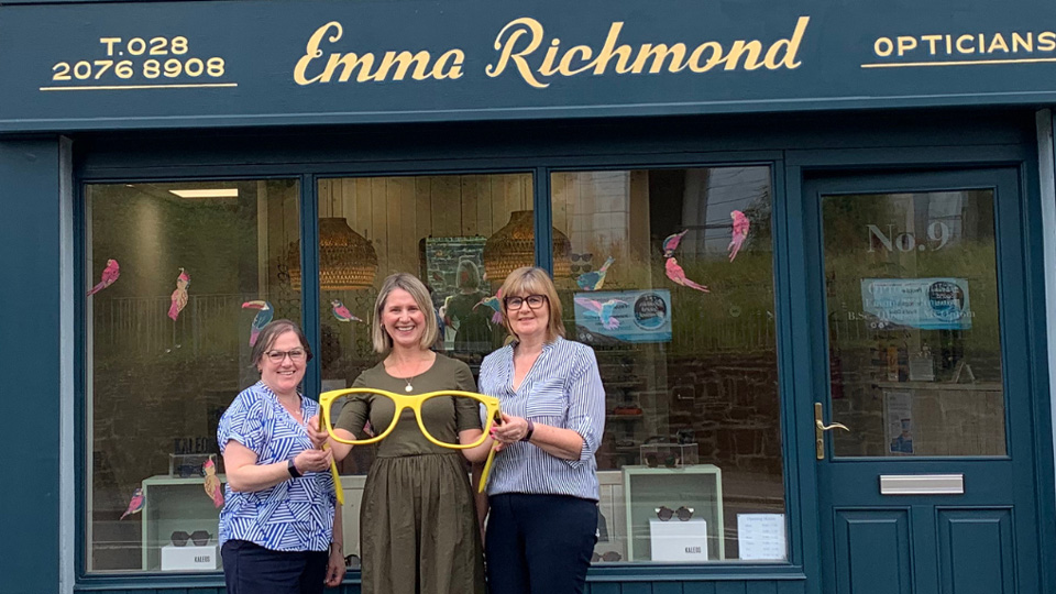 Three women stand outside an optometry practice holding a large pair of yellow glasses