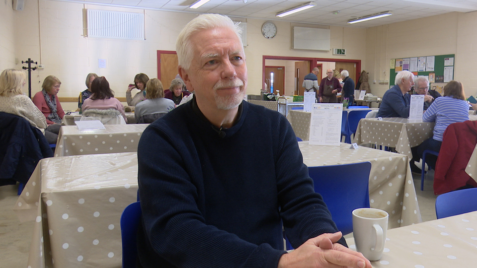 Professor David Thompson wears a navy jumper and sits in a community centre at a table covered in a beige and white dotted cloth. A cup of tea rests beside him. He smiles softly. There are people seated in groups around tables behind him