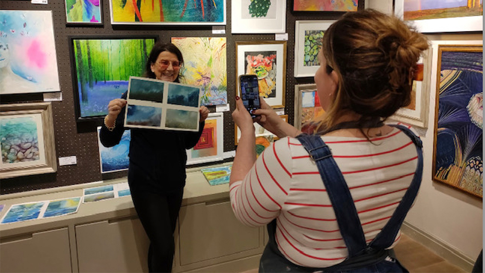 A photographer takes a picture of Francesca Marchetti who stands with a painting she has created against a backdrop of other paintings