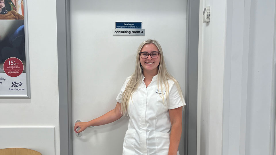 Kiera, wearing a white clinical uniform, smiles in a Boots Opticians testing room