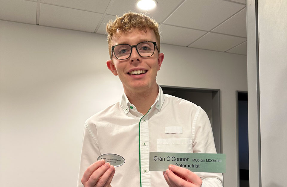 Oran holds up a badge and a door sign with his name and the title ‘Optometrist’