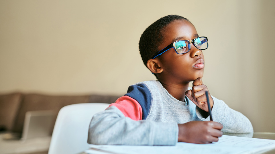 A boy wearing spectacles holds a pencil in his hand while reading an exercise book in front of him