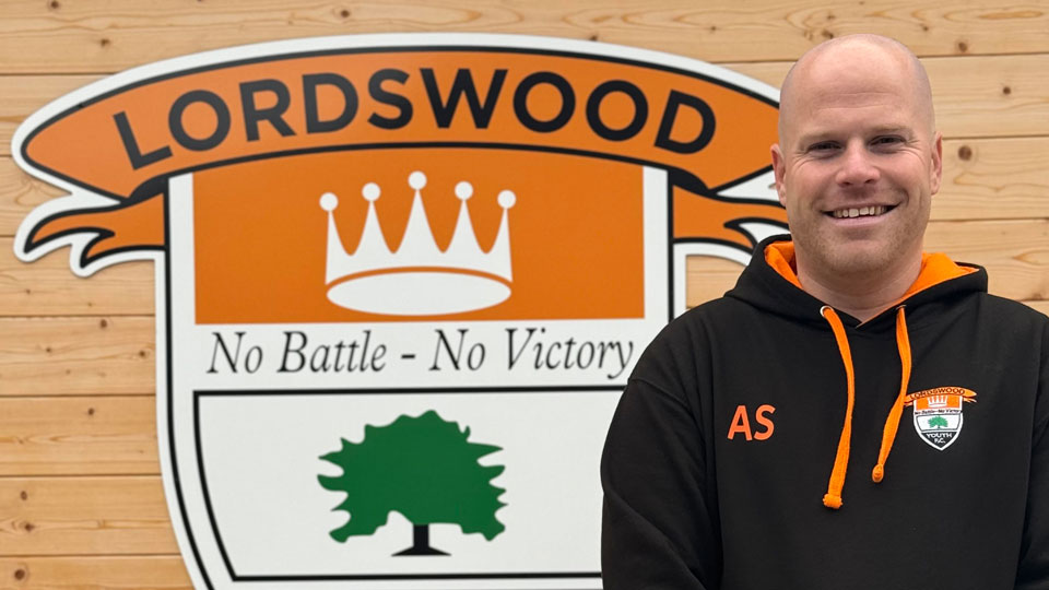 Ashley Smith stands by a large logo for the Lordswood Youth FC – the banner has a white crown on an orange background, a tree logo, and the motto: No battle – No victory. Ashley wears a black and orange hoody