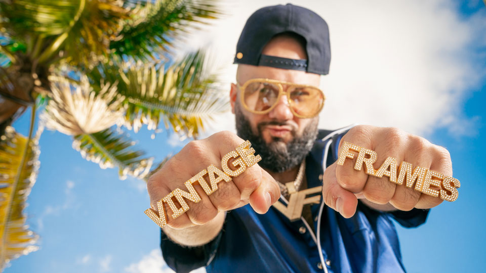 Corey wears rings that spell out ‘Vintage Frames’ in gold and diamante. Behind him is a bright blue sky and palm trees. He wears a sporty top, a gold chain with oversized ‘VF’ pendants and a backwards cap