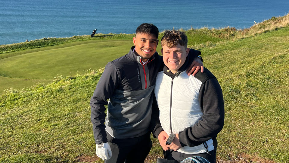 Zak has an arm around the shoulders of a friend. Both stand on a hilly golf course overlooking the sea