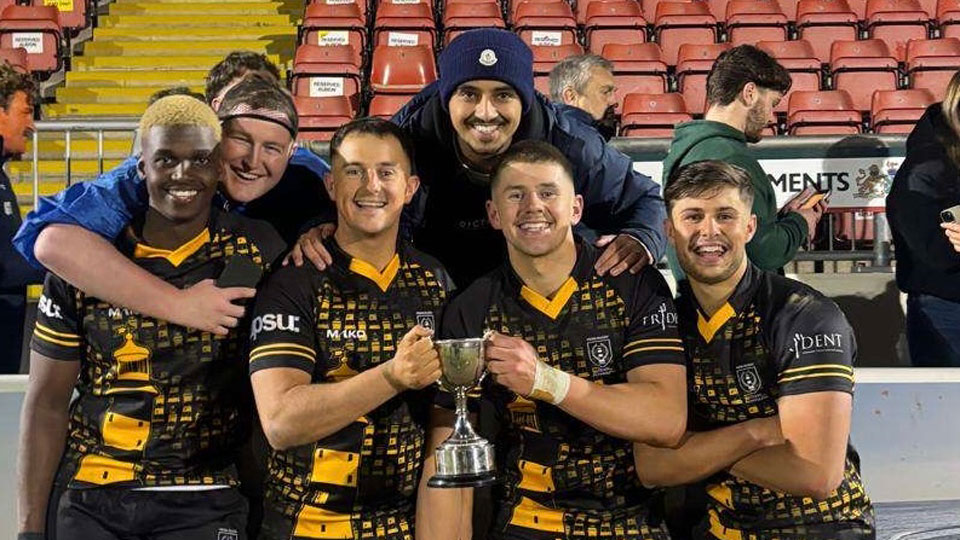 Four men in sports kit hold a trophy. Behind them, Zak and another friend lean over grinning for a celebratory photo