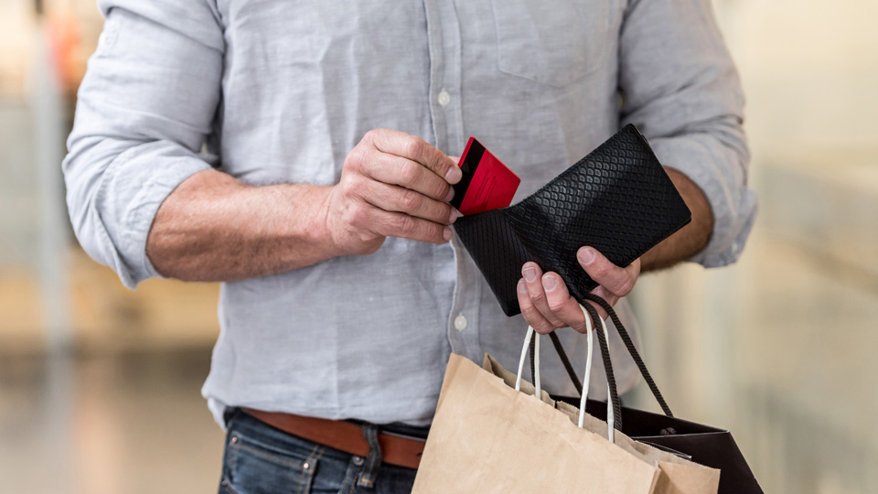 A man puts a credit card back into his wallet while holding shopping bags 