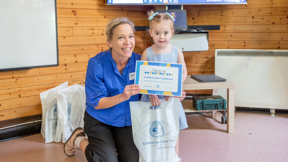 Lena, a support worker for CHECT, in a Royal blue blouse, kneels with Jessy who is holding a certificate recognising her as a CHECT champion
