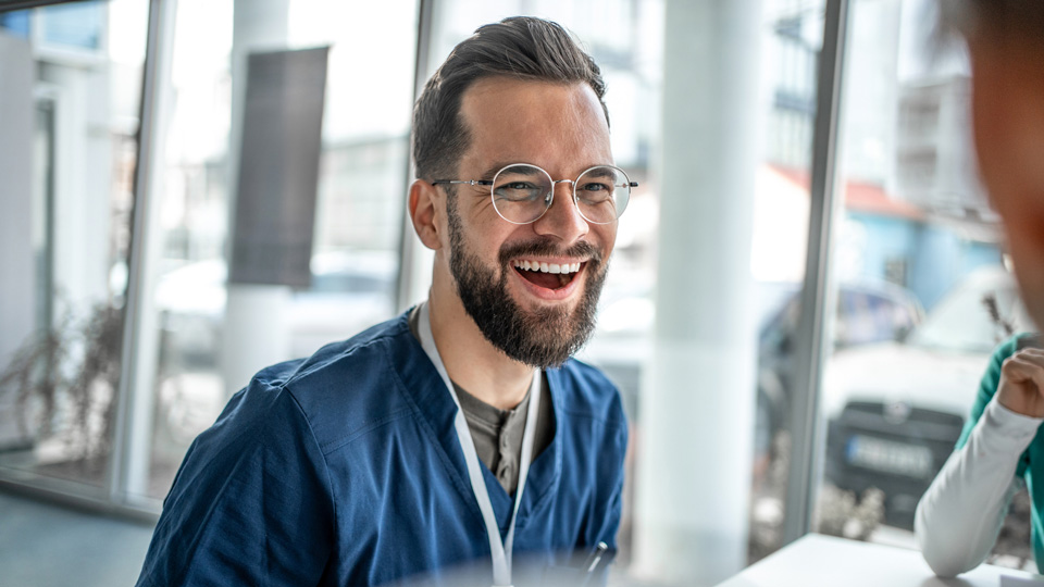 A male clinician wearing scrubs and glasses laughs
