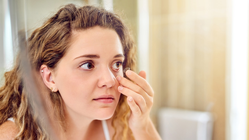 Female with blonde curly hair looking in a mirror with a contact lens on her finger preparing to put the contact lens in 