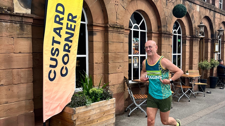 Ian wears running attire including a Samaritans branded top. He runs down a High Street with a large yellow banner reading Custard Corner