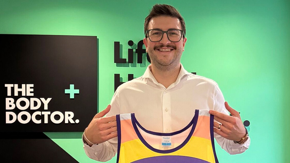 Sam stands in The Body Doctor offices holding up a running vest branded by the Pancreatic Cancer UK charity