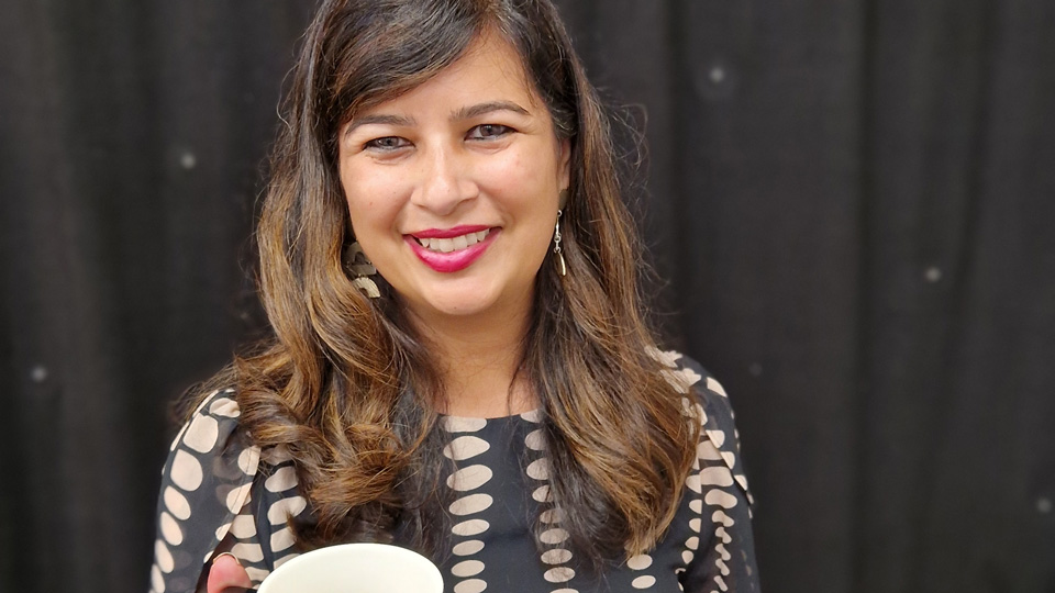 Sheena wears a black and white dress and pink lipstick, she holds a mug up to the camera like she is offering the viewer a coffee