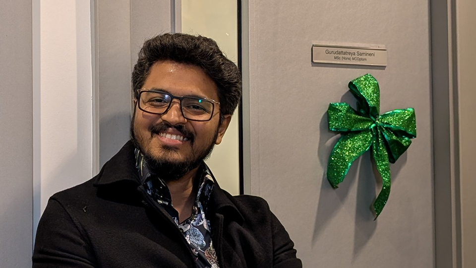 Guru is smiling with his arms folded whilst leaning against a wall