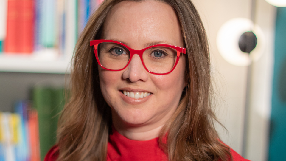Ceri Smith-Jaynes has brown curled hair that reaches her collar. She wears bright red frames with a slight cubic shape to the corners. The red matches the high-necked and long-sleeved red blouse she wears. Behind her are library bookshelves