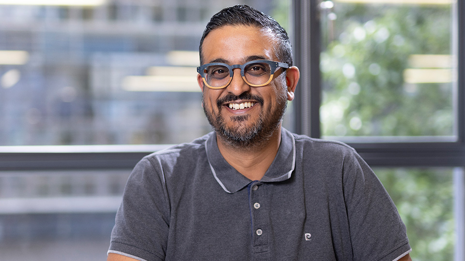 Ankur sits at a table smiling with hands crossed