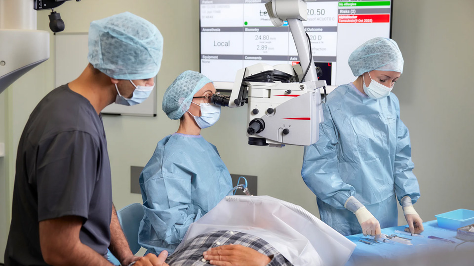 Three people standing in scrubs in an ophthalmology theatre around a table with a patient on it