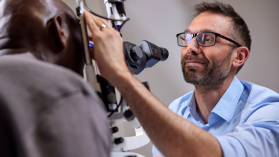 A man in a blue shirt sitting at a piece of optometry equipment to arm up testing a person’s sight