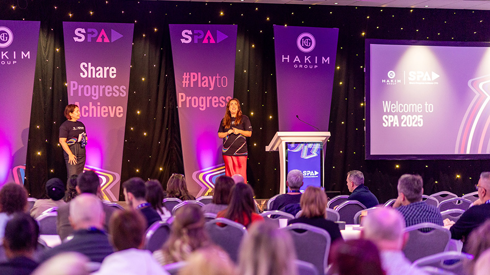 A conference speaker addresses an audience from the stage at the Hakim Group SPA 2025 event. Two presenters wearing SPA-branded T-shirts stand under purple lighting, with large banners reading “Share Progress Achieve” and “#PlaytoProgress” behind them. A podium sits beside a large screen displaying “Welcome to SPA 2025,” while attendees are seated facing the stage