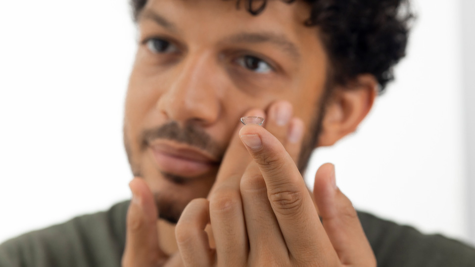 Close up view of a young male adult holding a contact lens on his finger. He is out of focus and the contact lens is in focus.
