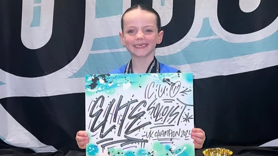 Harper is seated behind a row of trophies for dance competitions and holds a sign with the words ‘Elite’ and ‘Champion’