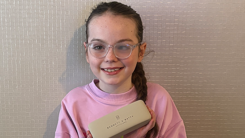 Harper wears spectacles with one frosted lens. She holds a spectacle case with the Bennett and Batty logo